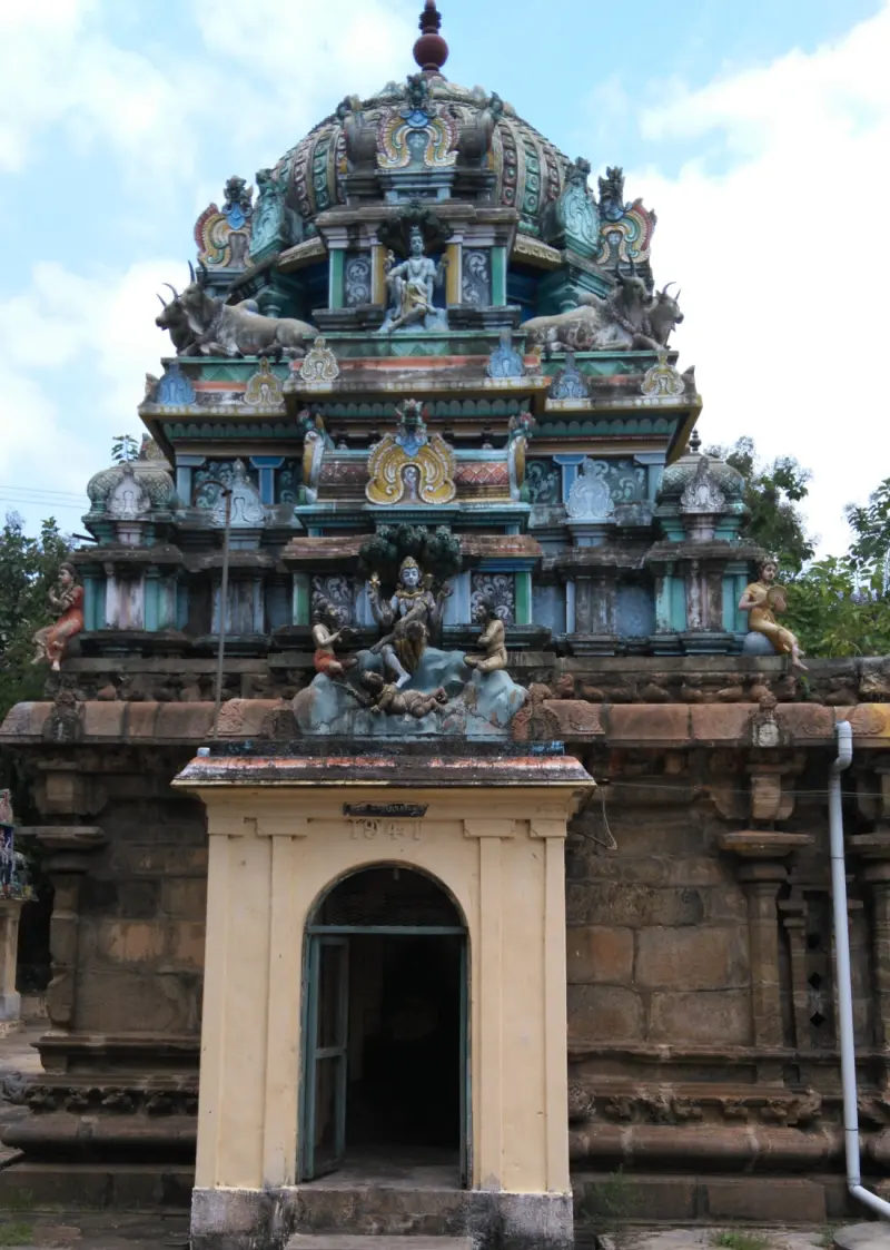A stone shrine under a tiered painted tower; a seated figure sits in a niche above a small yellow doorway.
