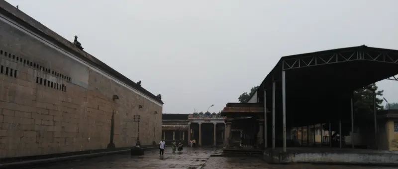 A tall stone wall on the left runs beside an open paved courtyard under a grey sky.