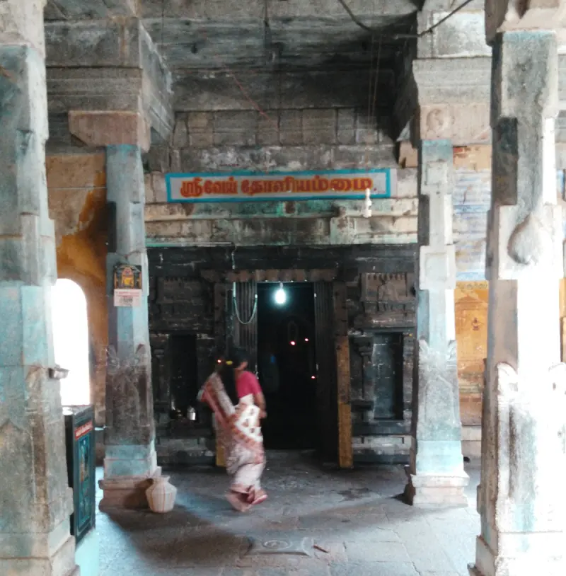 A shrine doorway between stone pillars with a Tamil name board above; a woman walks past in the corridor.