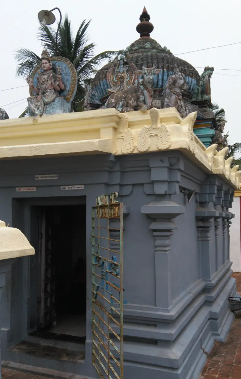 Side view of a grey shrine with two small name boards, a gate, and a seated goddess figure on the roof.