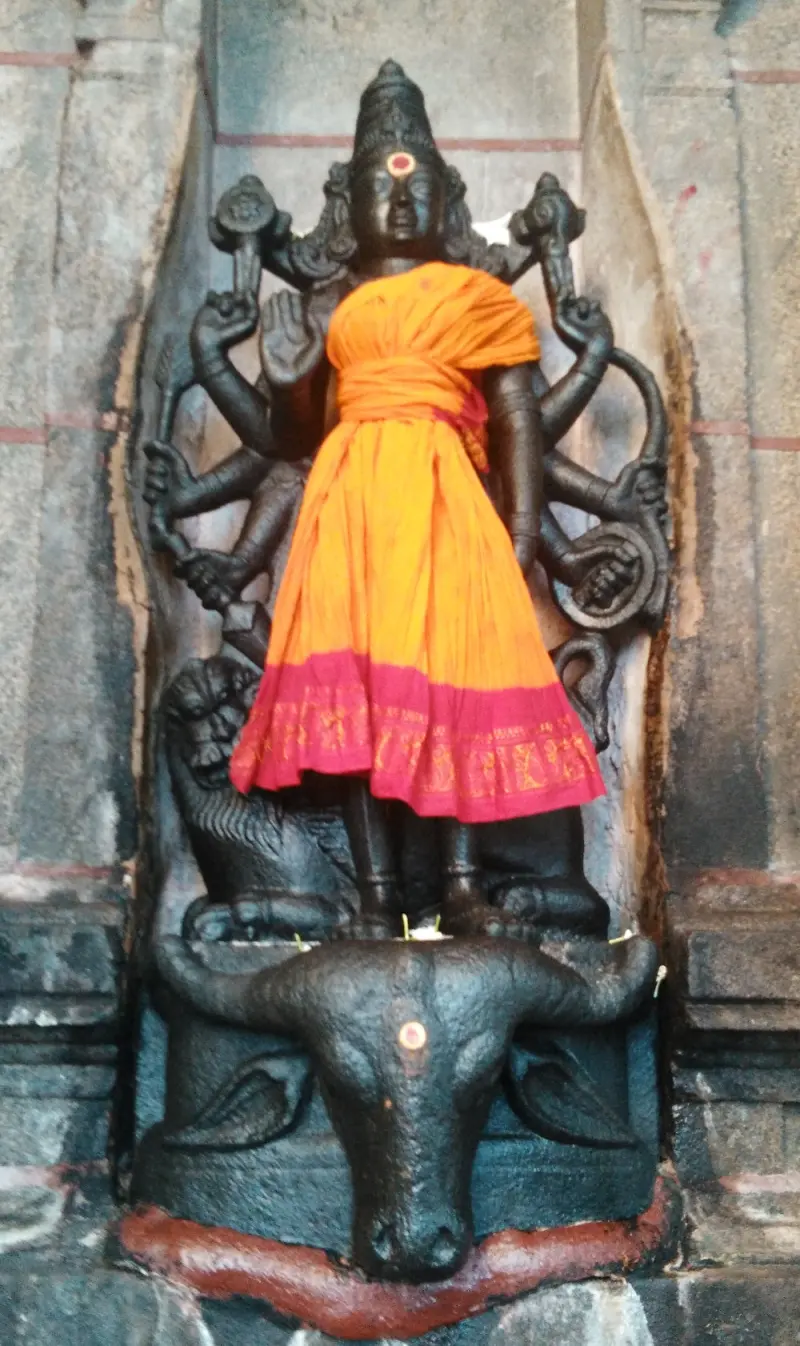 A many-armed black stone Durgai in an orange and pink cloth, with a lion behind her and a buffalo head below.