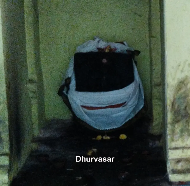 A dark stone idol wrapped in white cloth with flowers on top, with a label reading Dhurvasar.