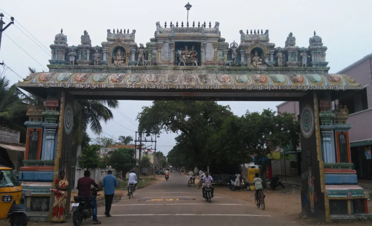 A painted gateway arch with small sculptures spans a village road, with riders and pedestrians beneath.