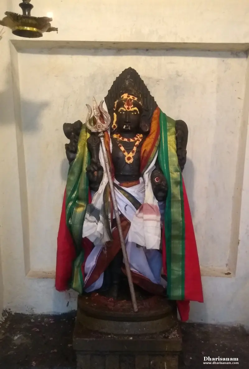 A dark idol in coloured shawls holding a silver trident, standing before a white wall