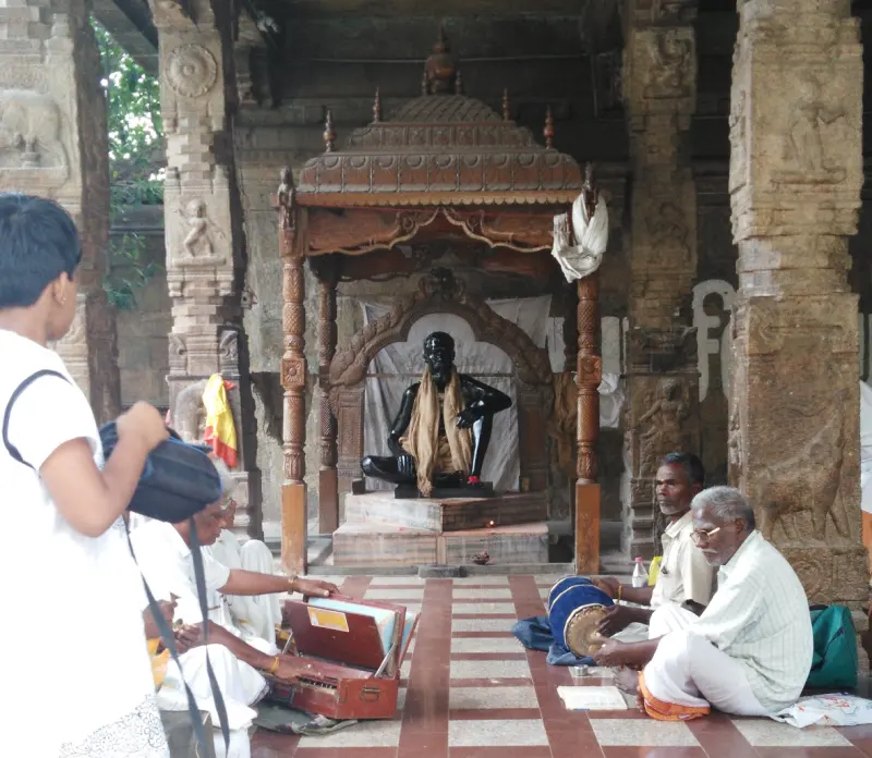 A dark idol under a wooden canopy in a pillared hall, with devotees seated with a harmonium