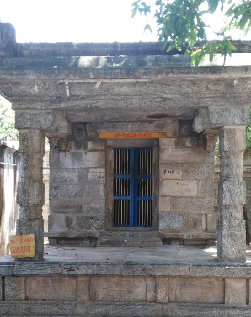 A small open stone shrine with a blue barred door and a Tamil name board above it.