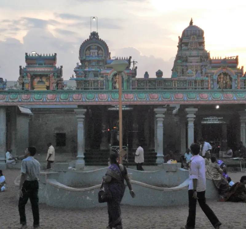 Visitors walk on a sandy courtyard in front of a hall whose roof carries painted figures and small towers