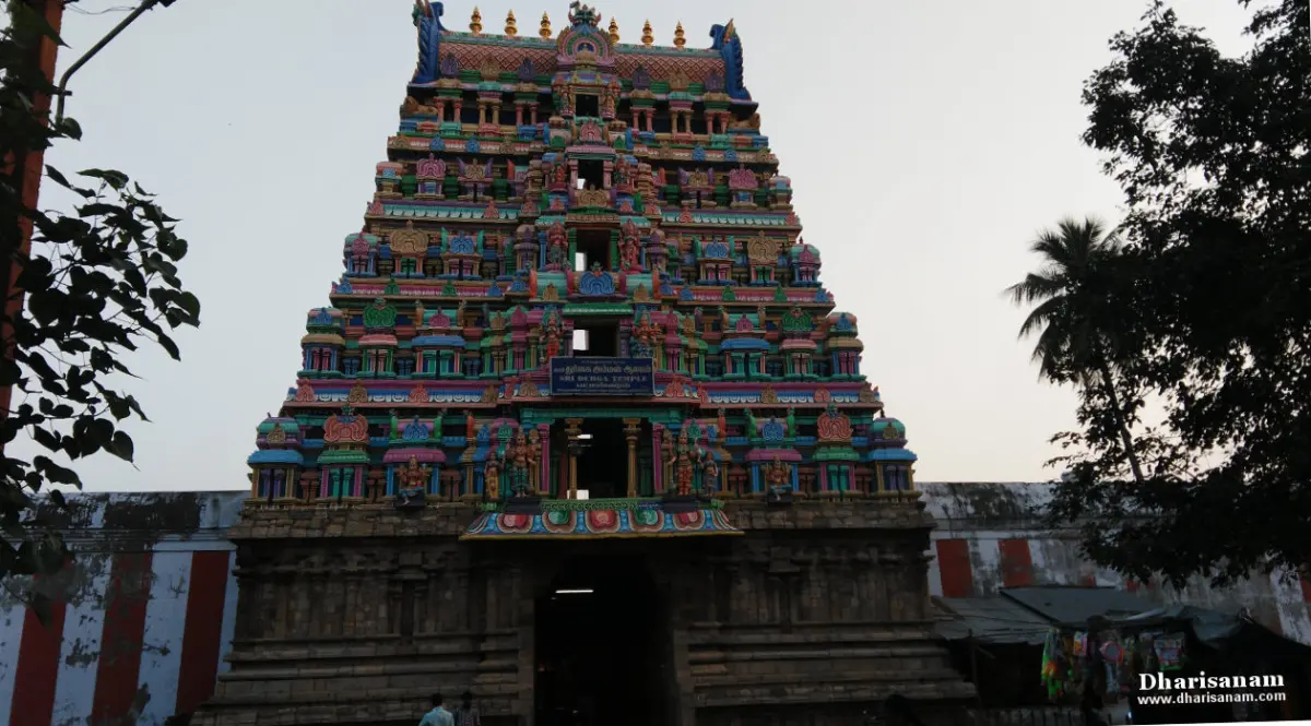 A tall tiered gopuram with a name board, a red and white striped wall beside it, and trees around