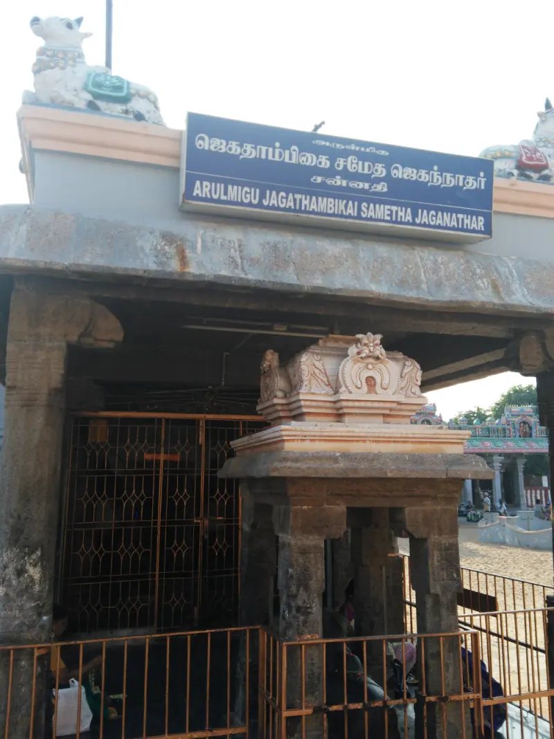 A shrine porch with a blue name board and white bulls on the roof, with a carved stone pedestal in front