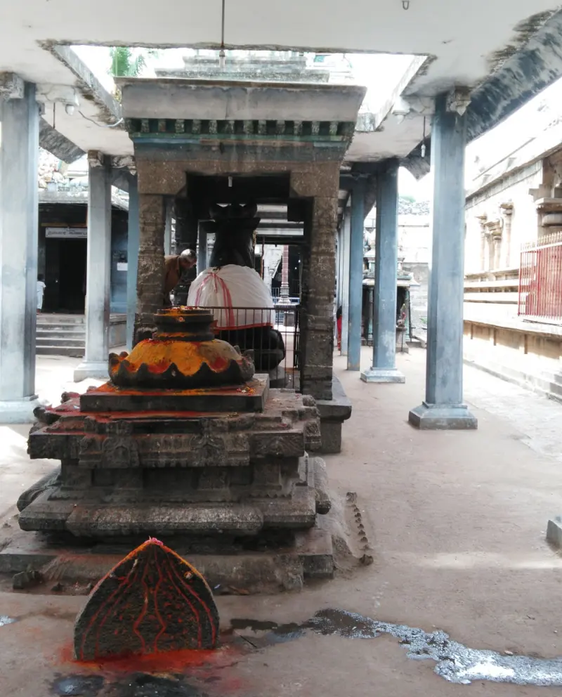 A stone pedestal with a lamp-like top and a Nandi seen beyond a small pillared frame, in a pillared corridor.
