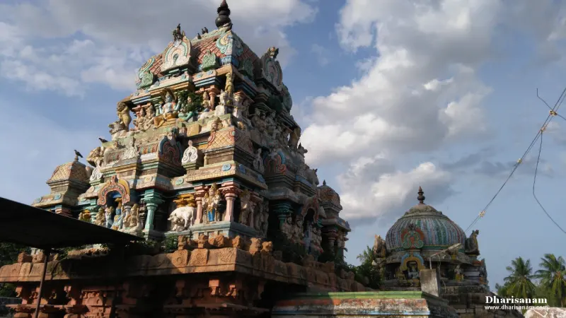 A sculpted tower rising against a cloudy sky, with a domed shrine to its right.