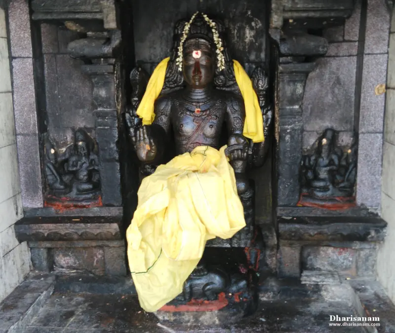 A dark stone idol draped in yellow cloth and a flower garland, seated in a niche with small carvings on both sides.