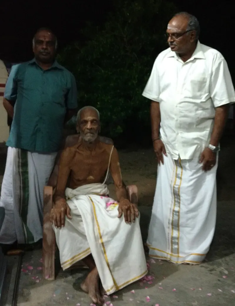 An elderly man in a white dhoti sits in a chair, with a man standing on each side