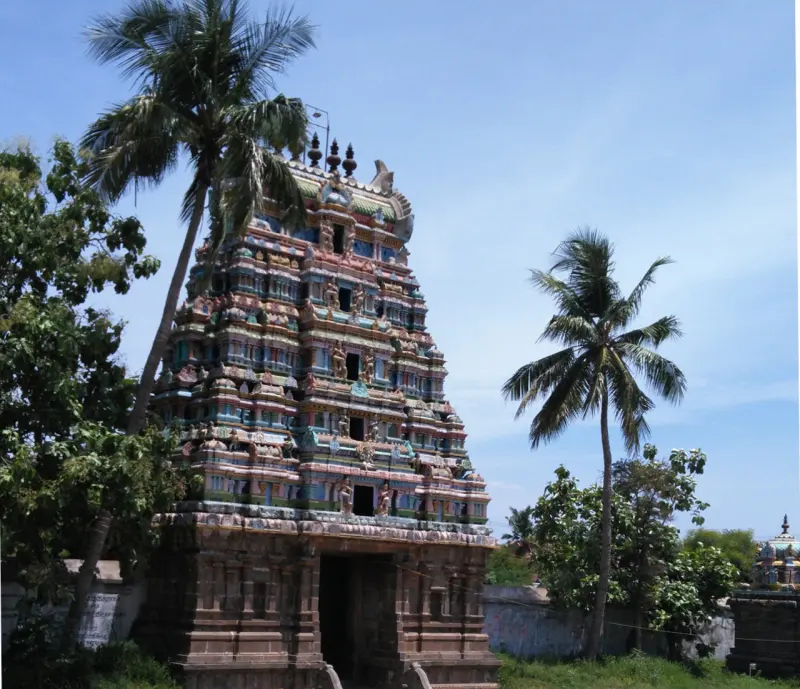 A colourful tiered tower on a stone base, with palm trees on either side and a wall in front.
