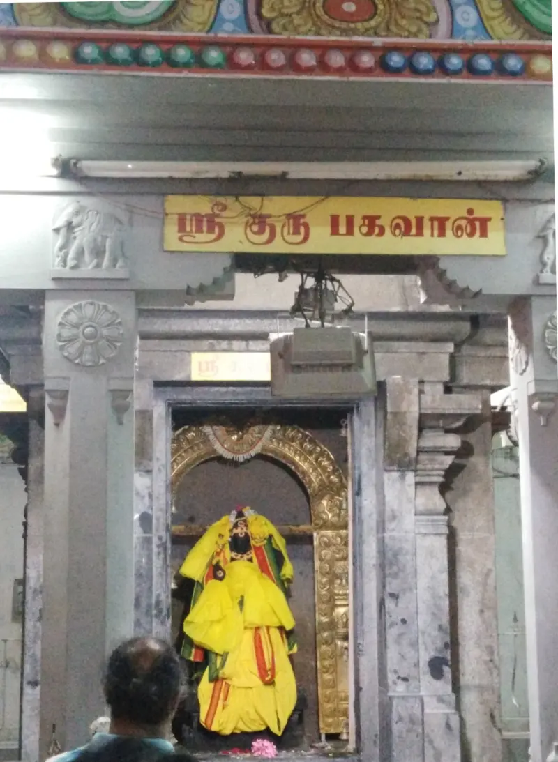 A dressed idol in yellow cloth inside a golden arch, under a yellow Tamil board reading Sri Guru Bhagwan