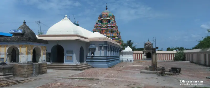 A wide paved courtyard with white domed roofs, a small arched stone shrine at left and a painted tower beyond