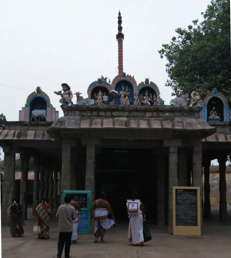 Devotees stand at a pillared mandapam with small figures on its roof and a flagpole behind