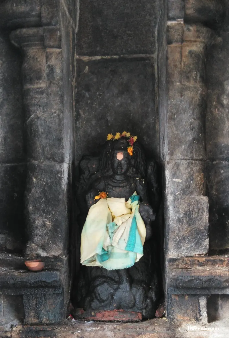 A dark seated idol with a flower crown and a light cloth sits in a deep stone niche, with a clay lamp beside it