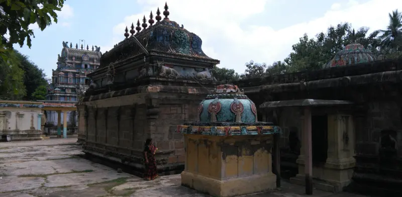 Two stone shrines with painted domes in a paved courtyard; a tower rises behind and a woman stands at left.