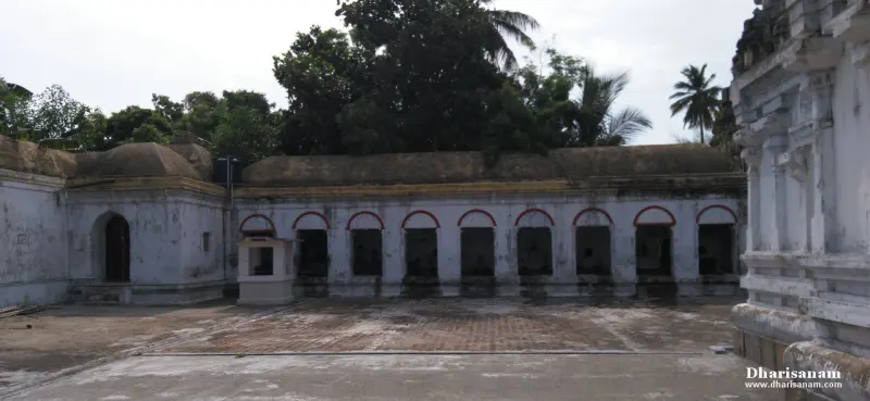 A long corridor with red-arched openings faces a paved courtyard, with trees rising behind its roof