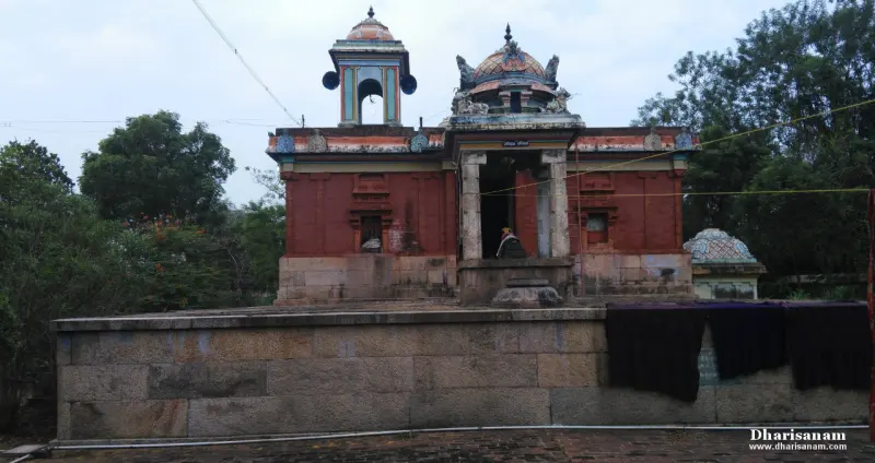 A red-walled temple with a small mandapam and a bell tower, behind a long stone wall and under tall trees.