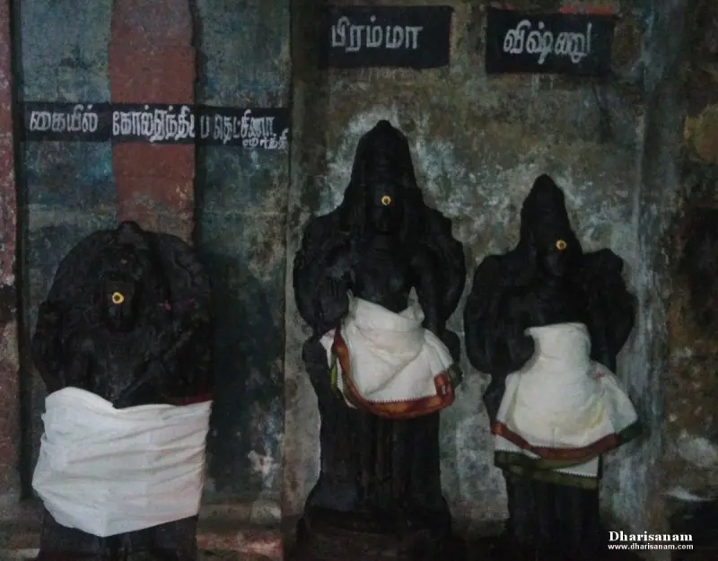 Three black stone idols in white cloth stand against a wall, Brahma and Vishnu name boards above two