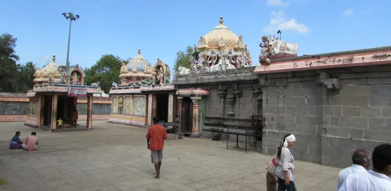 A paved courtyard with several shrines with domed roofs, a stone wall to the right, and visitors walking.