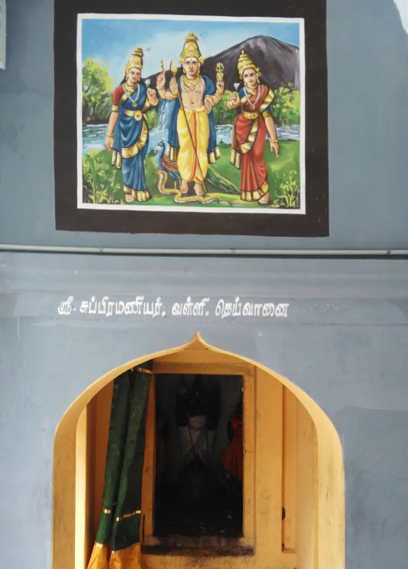 An arched shrine doorway below a painting of Murugan between two goddesses and a Tamil name line