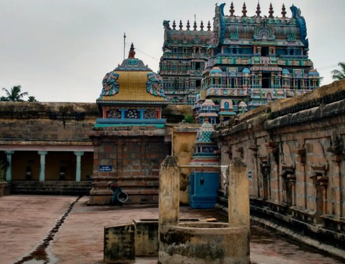 A colourful tower rises behind a golden-domed shrine and stone corridor walls, with a stone pillar base in front.