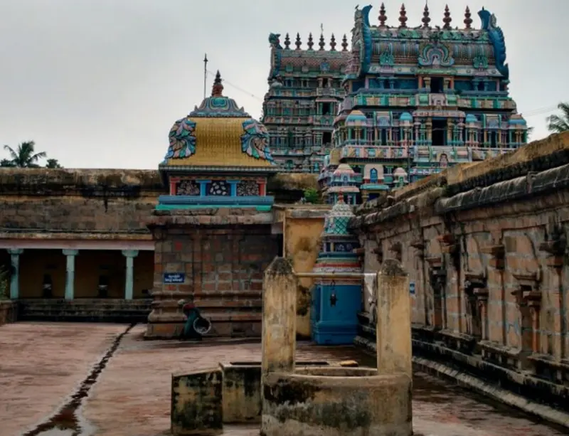 A colourful tower rises behind a golden-domed shrine and stone corridor walls, with a stone pillar base in front.