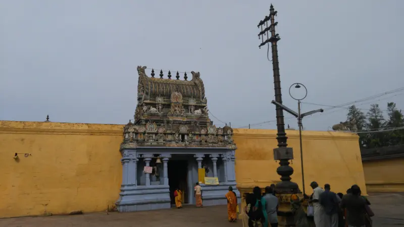A small painted gateway in a yellow wall with devotees and a tall flagpole at the right.