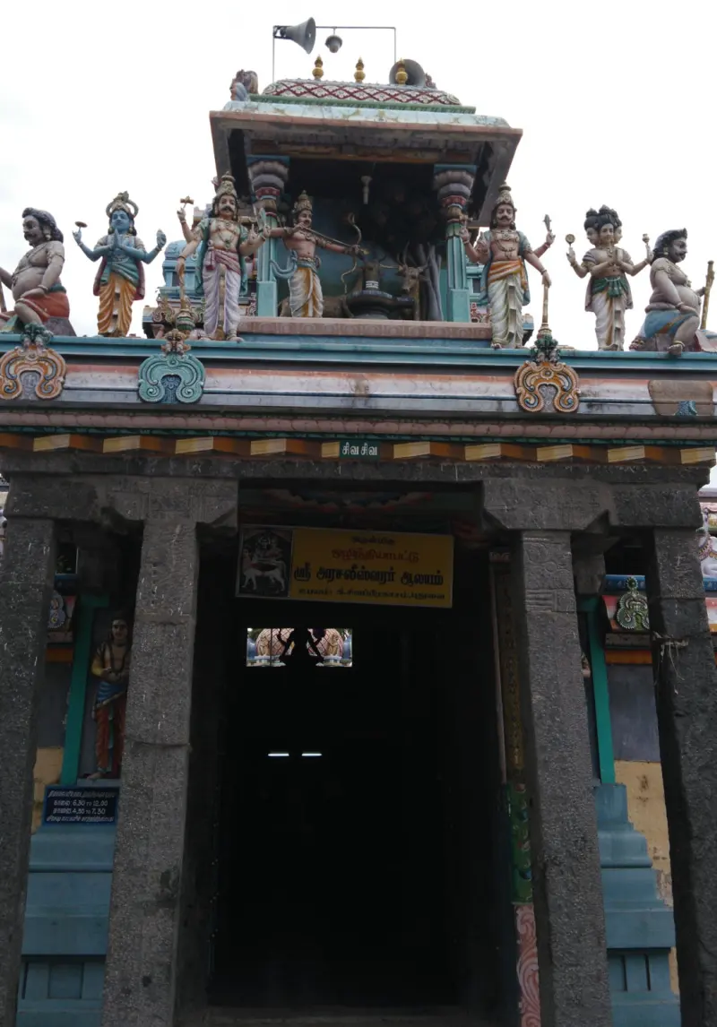 A stone gateway with a name board in Tamil and a small tower above it with painted figures of deities and a deer.