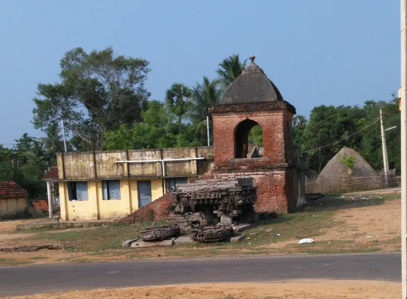 Carved stone wheels and remains of a car beside a brick tower, near a road