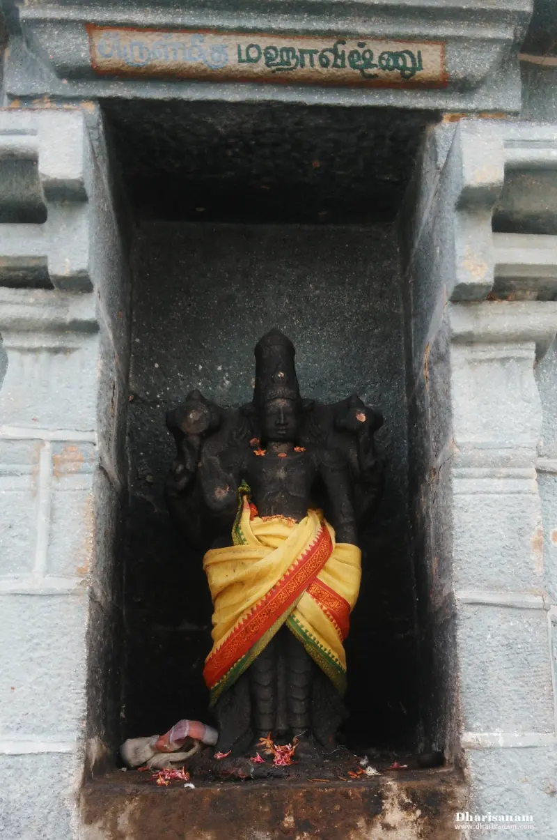 A standing four-armed black stone idol in a yellow and red cloth, with a Tamil name board above the niche
