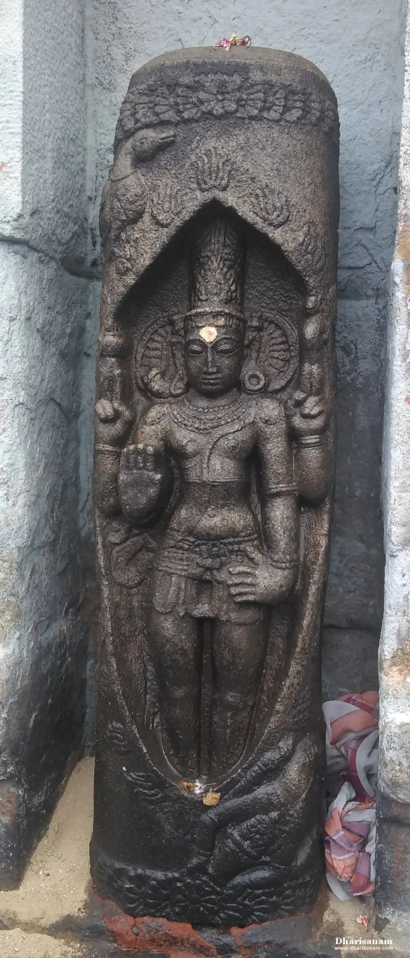Dark stone carving of a crowned four-armed deity standing in a niche