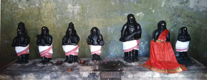 Seven small black idols in white cloth, one holding a lute, with a red-clad idol at the right