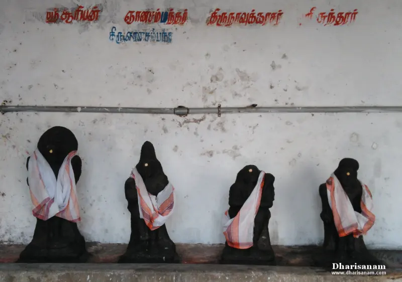 Four black stone idols wrapped in cloth stand in a row beneath Tamil name labels painted on a white wall