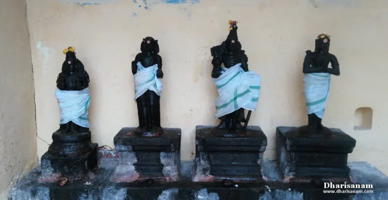 Four dark idols wrapped in white cloth stand in a line against a yellow wall, each on its own stone base