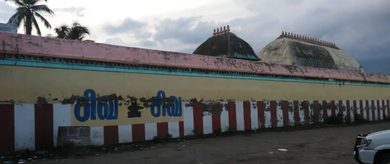 A long compound wall with red and white stripes and large blue Tamil lettering; two domed roofs rise behind it.