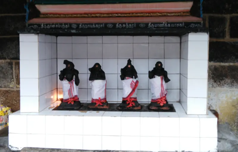Four small dark figures in a row, dressed in white and red cloth, inside a white-tiled shrine.