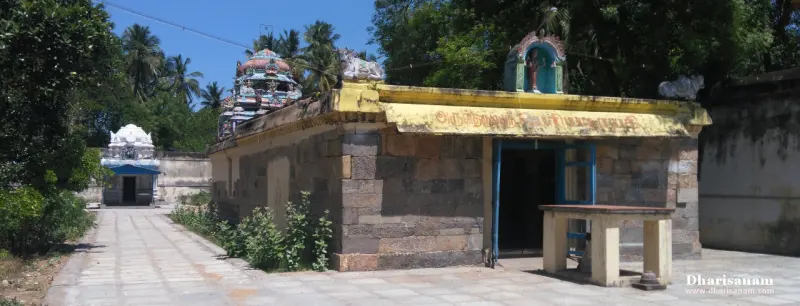 A stone shrine with a yellow signboard on its roof edge beside a paved path, with towers behind
