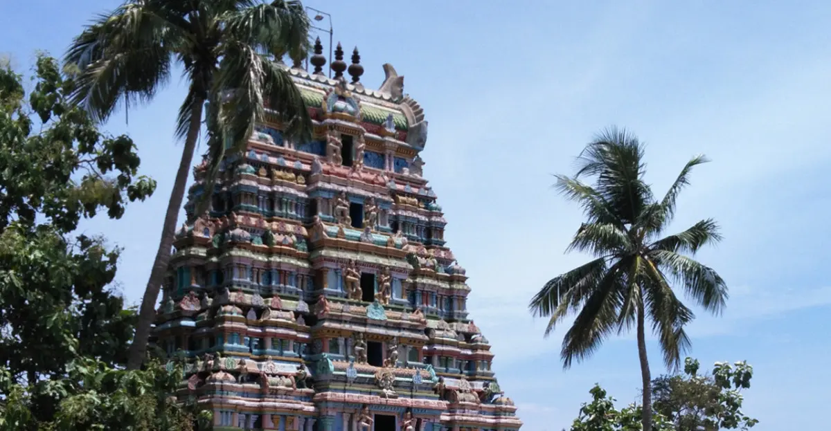 A tall tiered gopuram painted in many colours, with figures on each level, between two palm trees.
