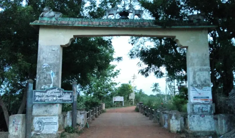 A weathered arch with a board on its left spans a red earth road lined with trees.