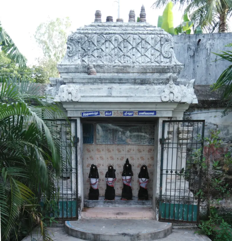 A small white shrine with four dark figures wearing garlands and cloths, behind an iron gate