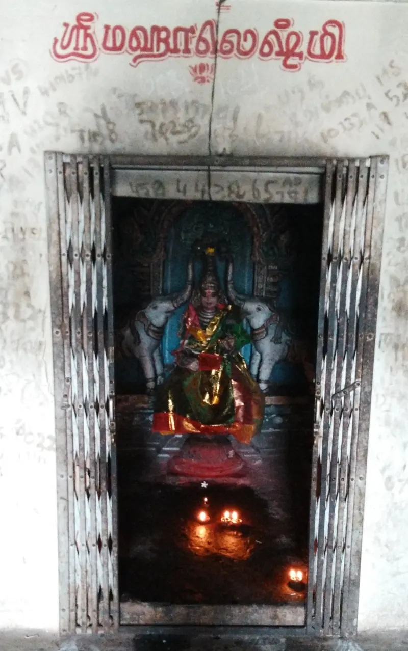 A seated figure in a green and gold sari with two elephants beside her, behind a folding iron gate and lit lamps.