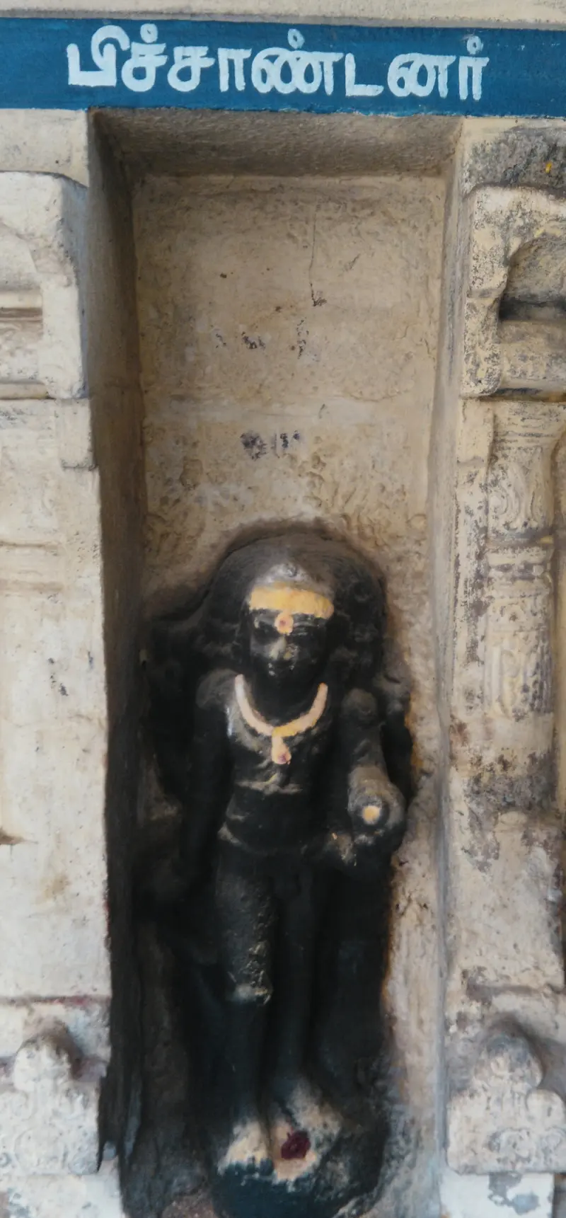 A dark standing idol with sacred ash marks under a blue name board