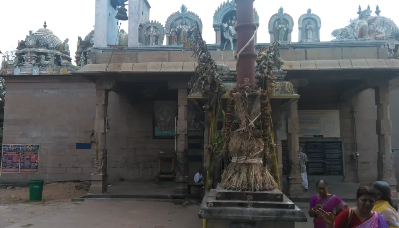 A flagstaff base wrapped in dry grass in front of a stone mandapam with painted figures along its roof