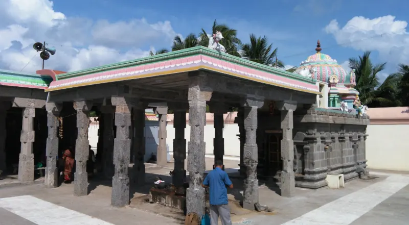 An open stone mandapam with a visitor standing in it and a colourful shrine dome beside