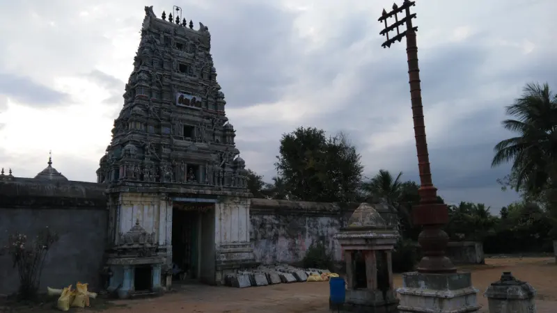A carved gopuram beside a tall flagstaff, with palm trees behind the compound wall