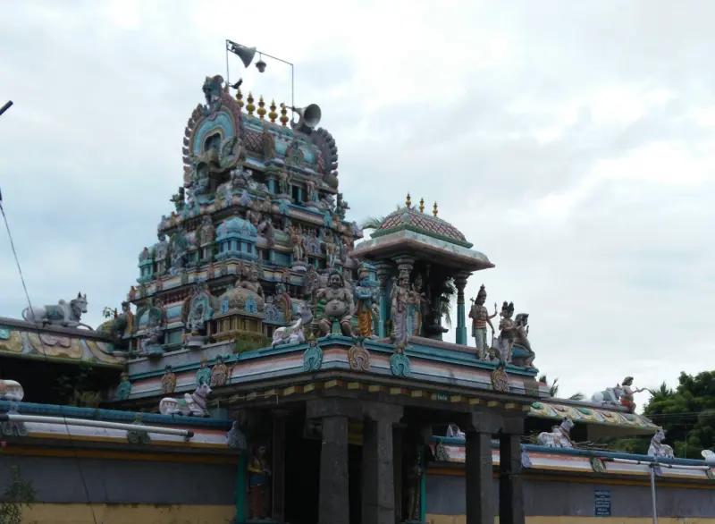 A small painted gopuram with many sculpted figures above a pillared gateway, seen under a cloudy sky.